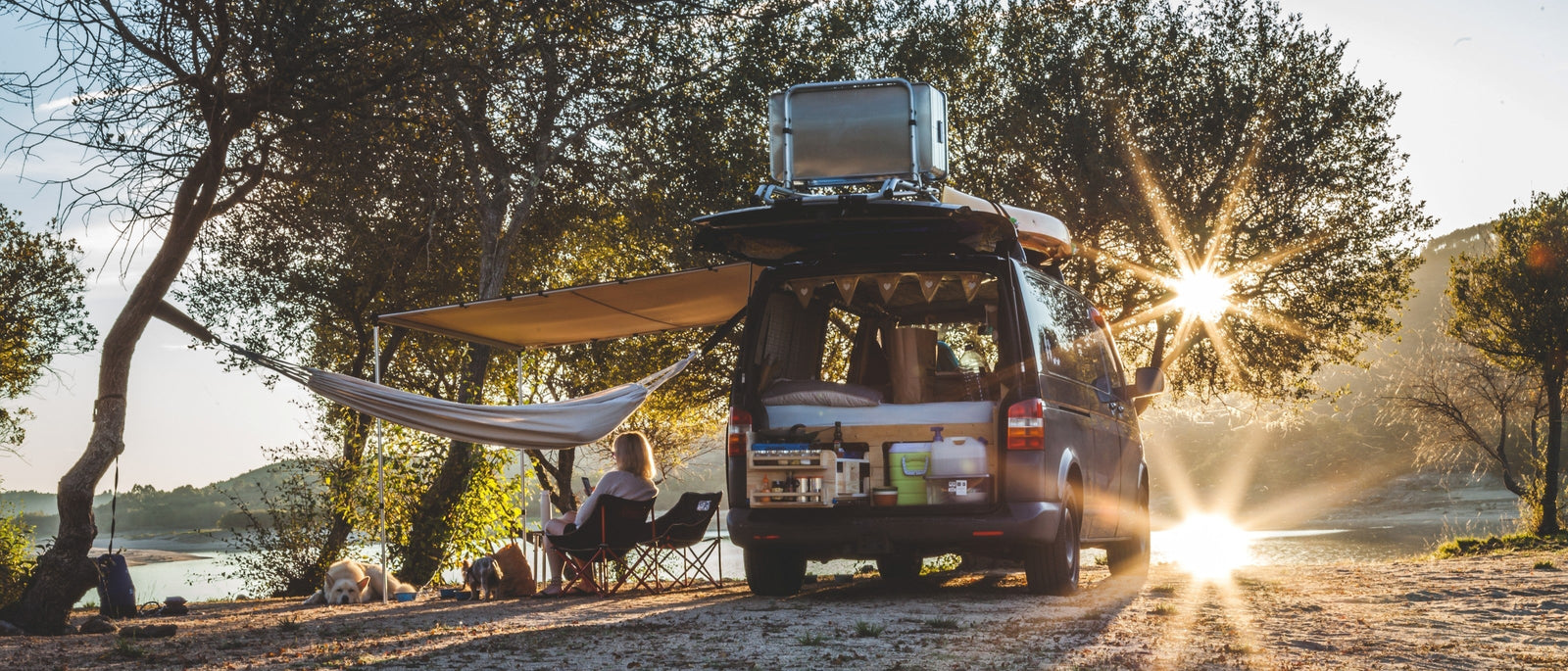Camper van with a side awning extended, a hammock strung between trees and two dogs relaxing beside it at golden hour, parked on a dusty track above a lake.