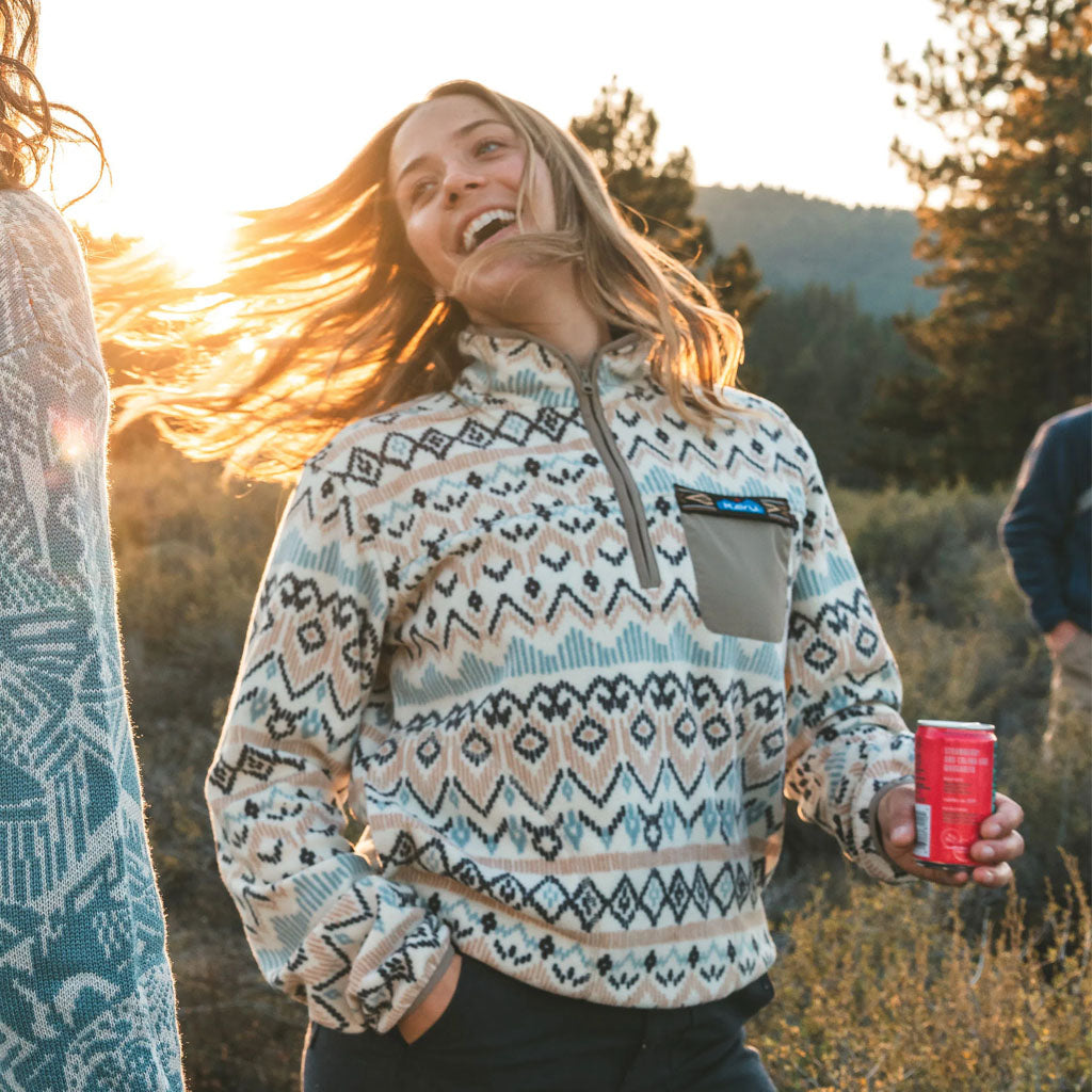Woman wearing a Kavu patterned fleece at sunset in mountains, holding a drink.