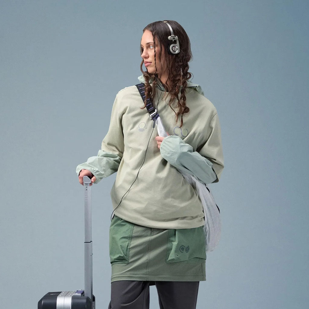 Woman wearing Purple Mountain Observatory hoodie and cargo shorts with backpack and headphones, holding luggage handle.
