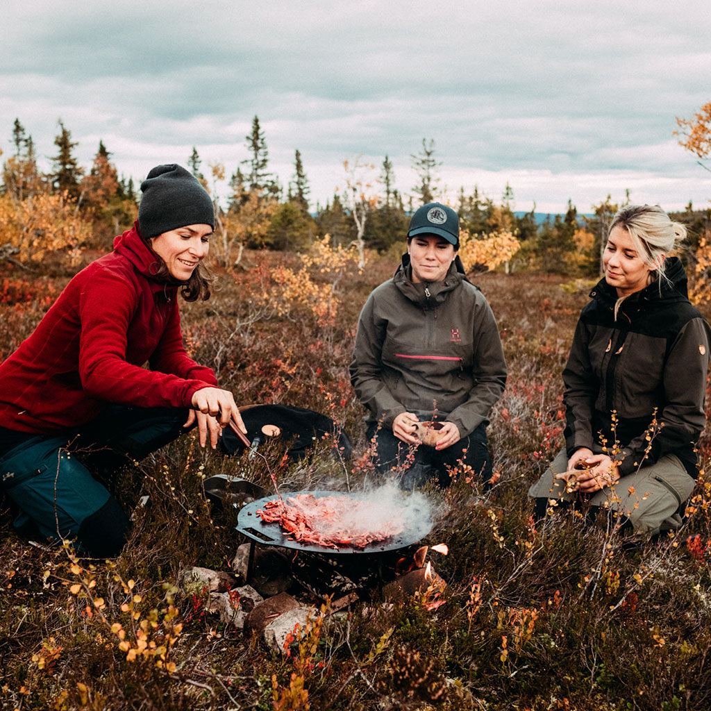 Three people cooking over a campfire in autumn woodland with coniferous trees and golden shrubs in the background.
