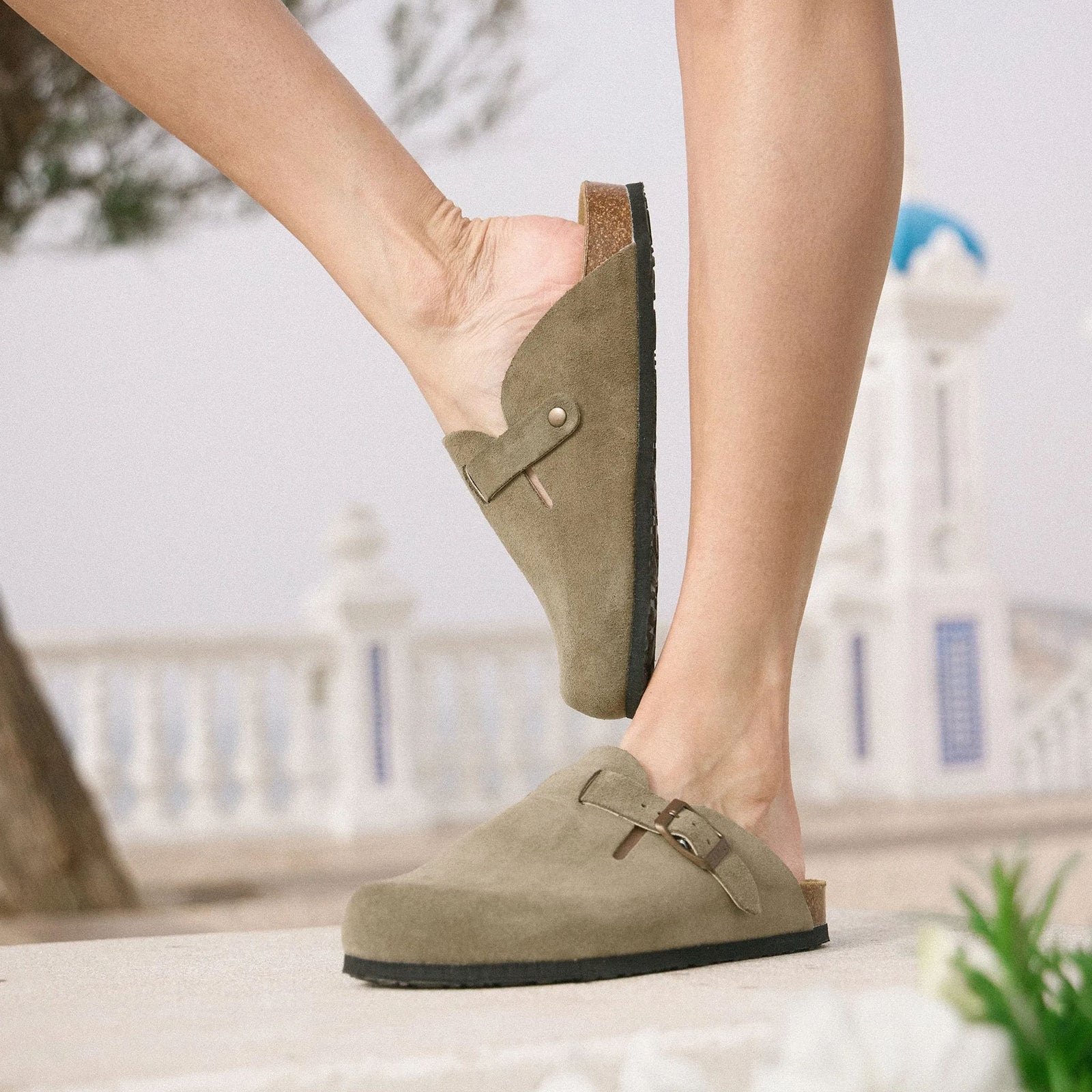 Plakton women's suede clogs with buckle detail, worn outdoors with coastal architecture in background.