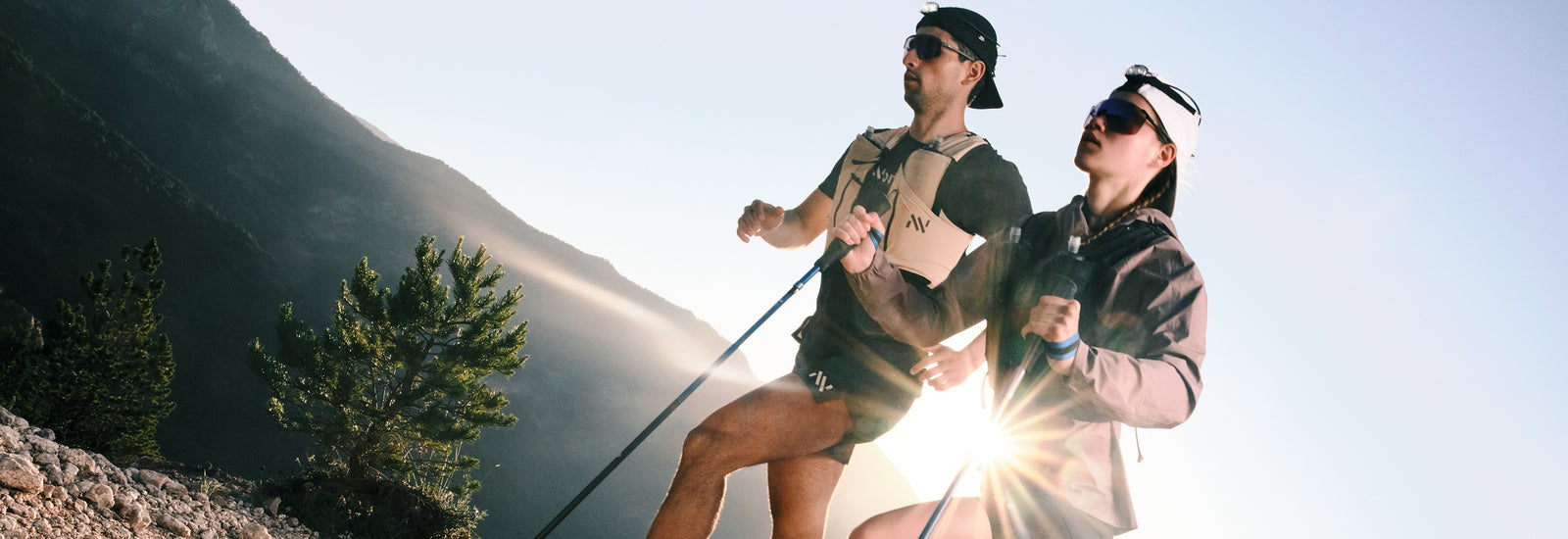 Two hikers in mountain sunlight wearing Nnorma clothing and caps while trekking with poles.