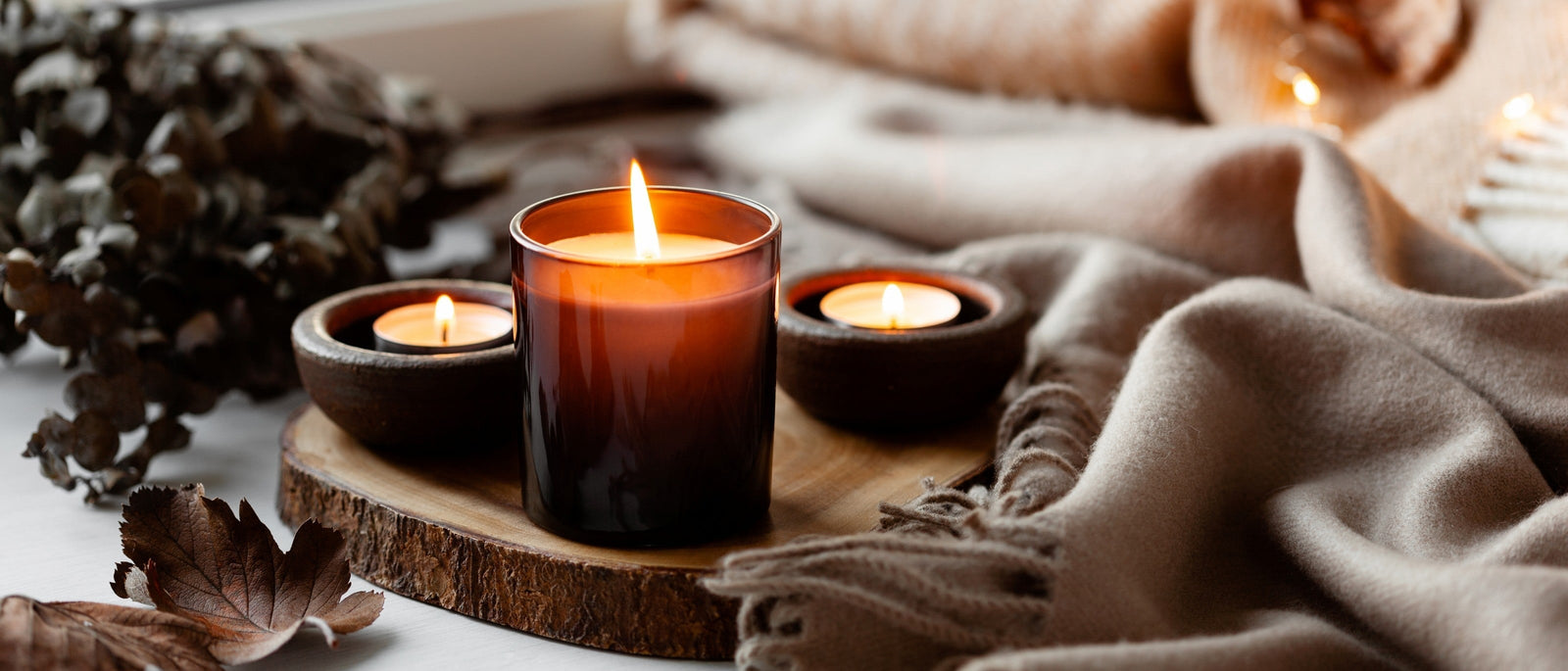 Multiple lit candles of varying sizes on wooden tray with soft blankets, tree bark and autumn leaves.