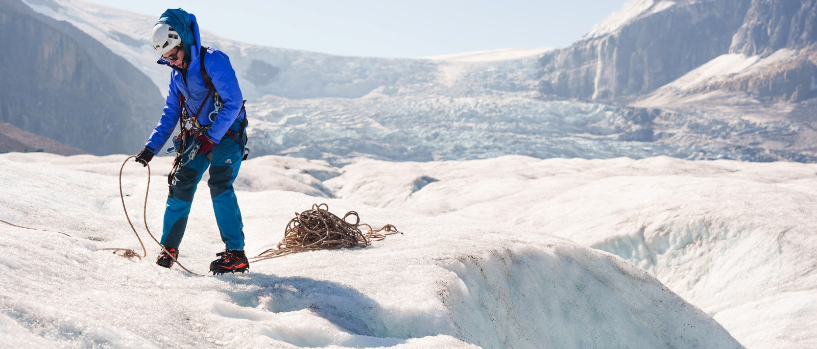 Climber in blue and teal stands on white glacier surface coiling rope with massive white icefall and mountain cirque behind.