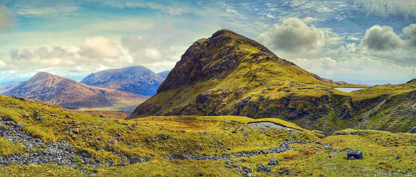 Marsco mountain on Skye with the Red Cuillin peaks behind, showing steep grassy slopes under partly cloudy skies, viewed from the approach to Bla Bheinn. Photo by Ellie Clewlow