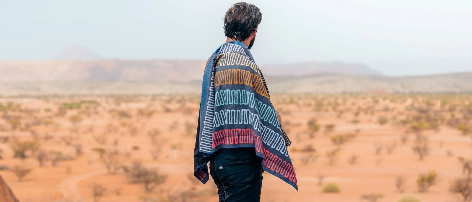 A man wrapped in a striped Nomadix Ultralight Towel in the Switchback Multi colourway looks out over a red desert landscape with distant hills