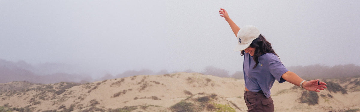 Woman in purple shirt and white cap balancing on a sandy dune in misty landscape.