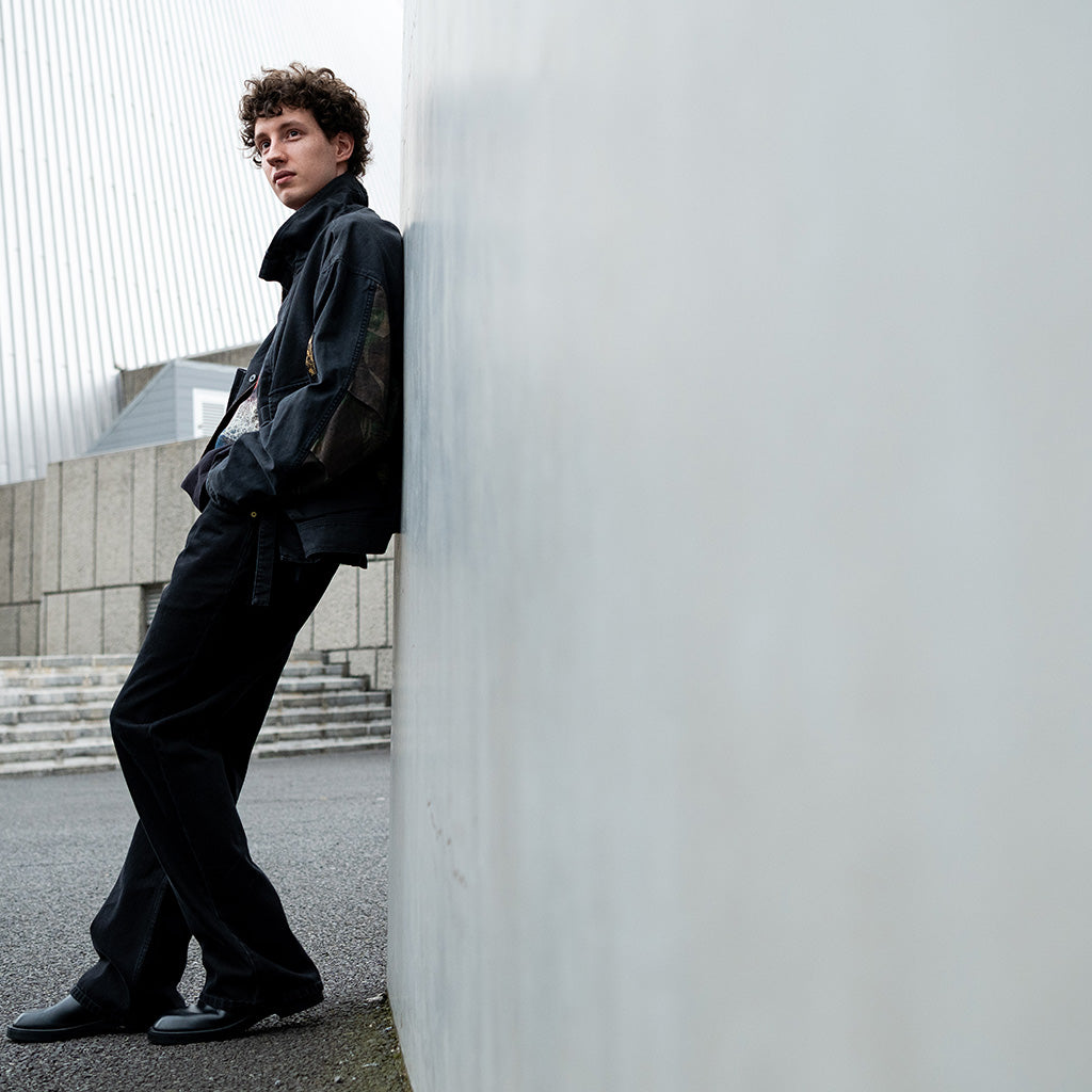 Person wearing dark denim jacket and jeans leaning against a white concrete wall in an urban setting.