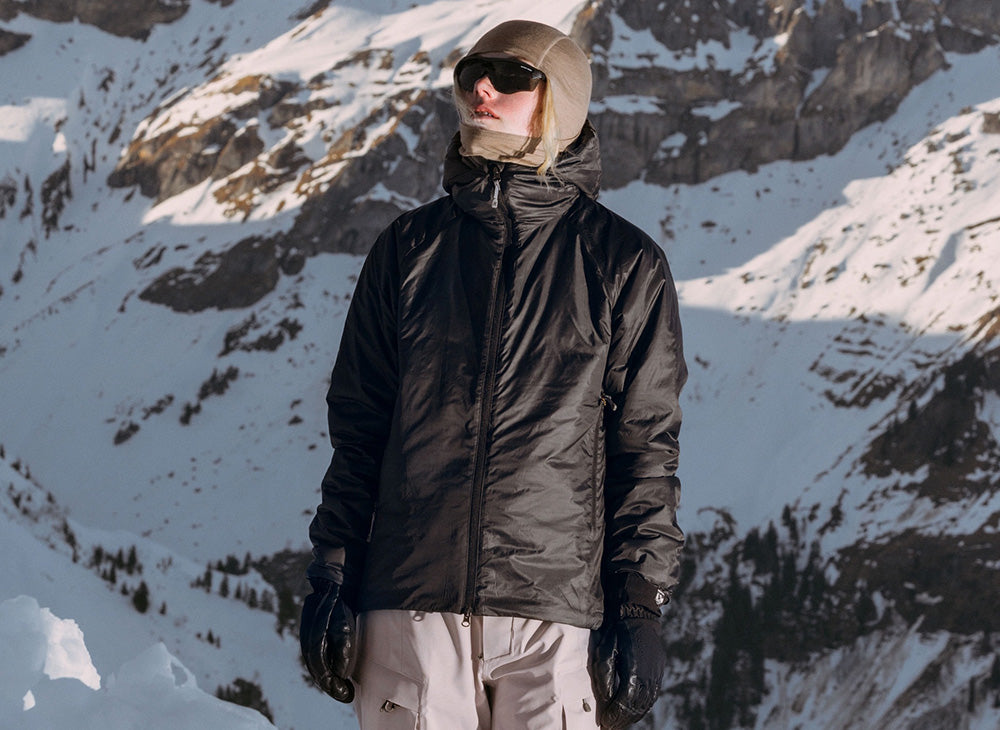 Houdini brand spotlight: a person in a black insulated jacket and white snow trousers on a snowy mountain slope.
