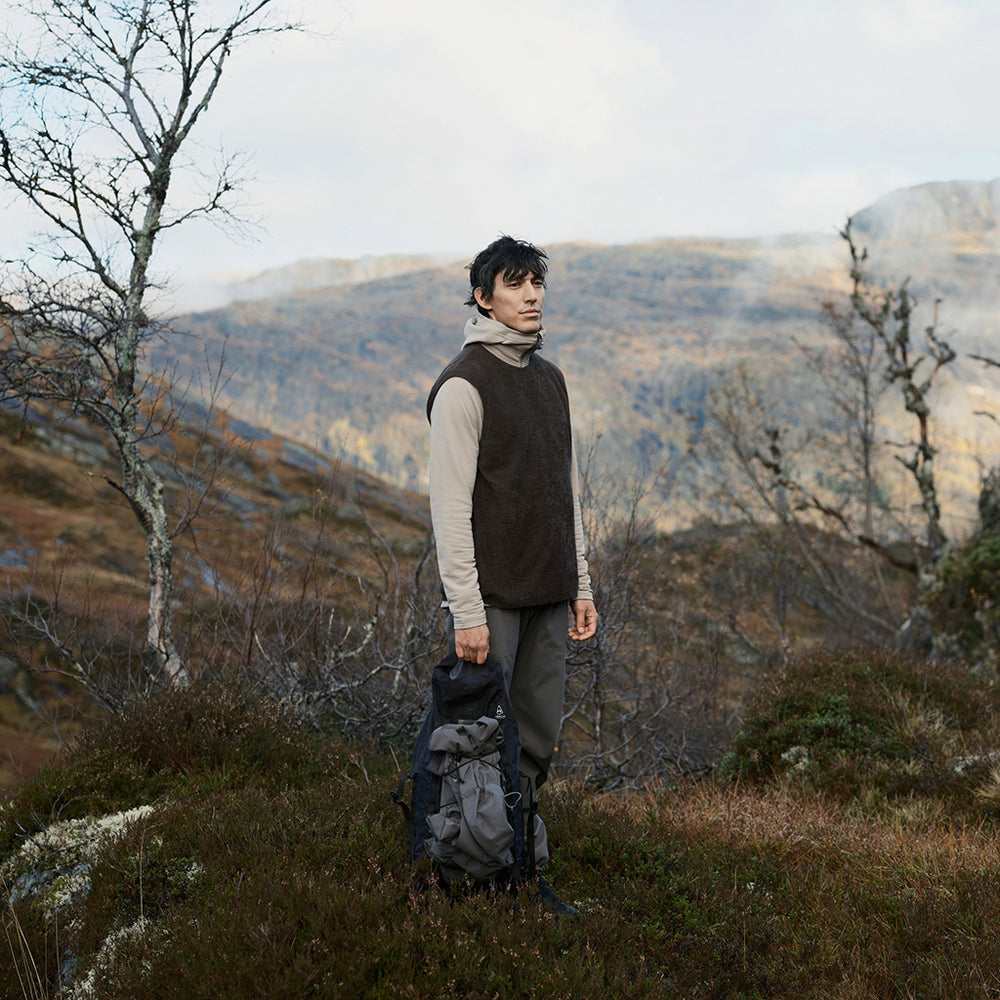 A hiker wearing a black gilet, grey hoodie and trousers stands on a hillside with a backpack, surrounded by bare trees