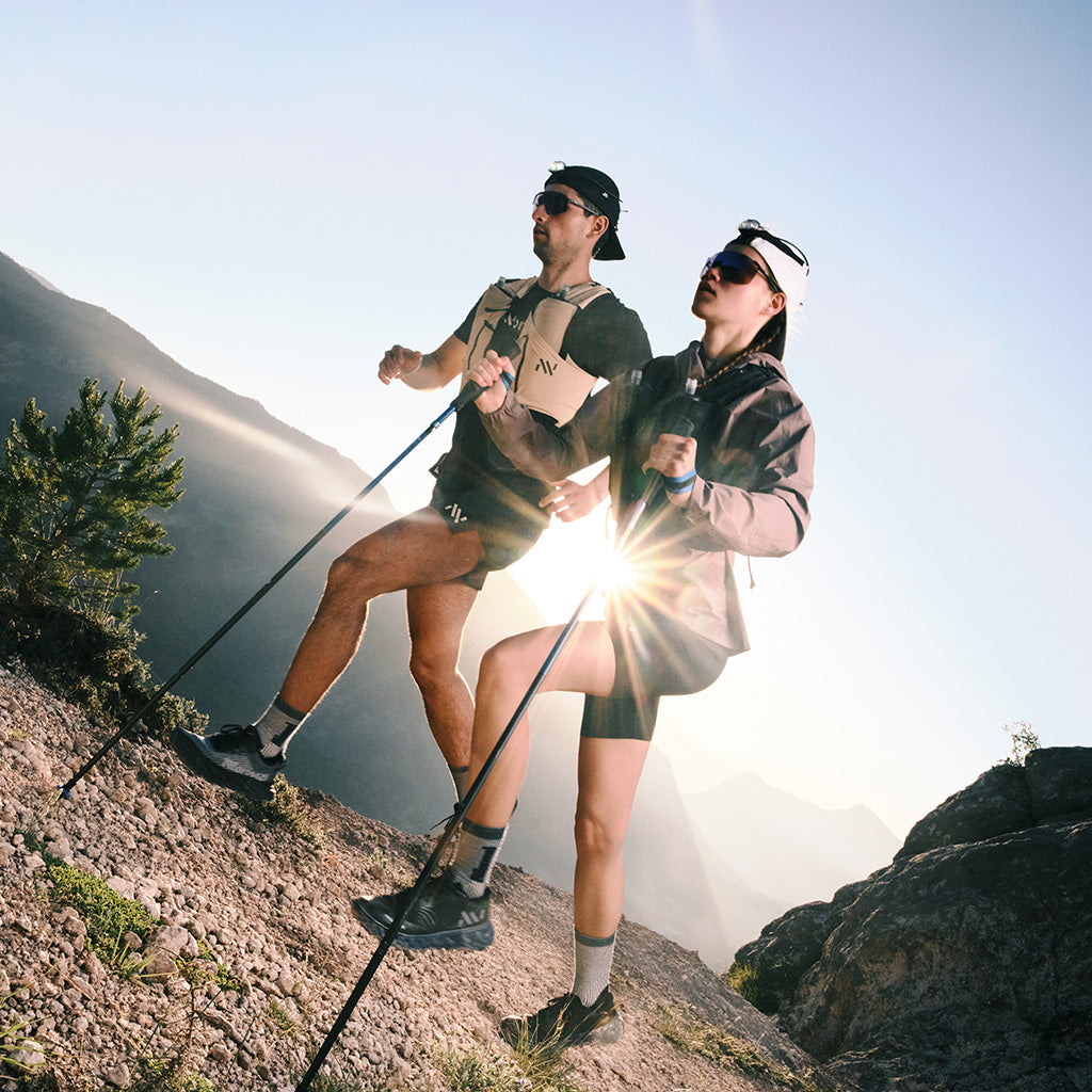 Two hikers running uphill on a mountain trail with trekking poles in bright sunlight.