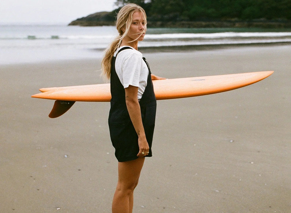 Surfer carrying an orange surfboard on a sandy beach, wearing a black wetsuit vest over a white t-shirt.