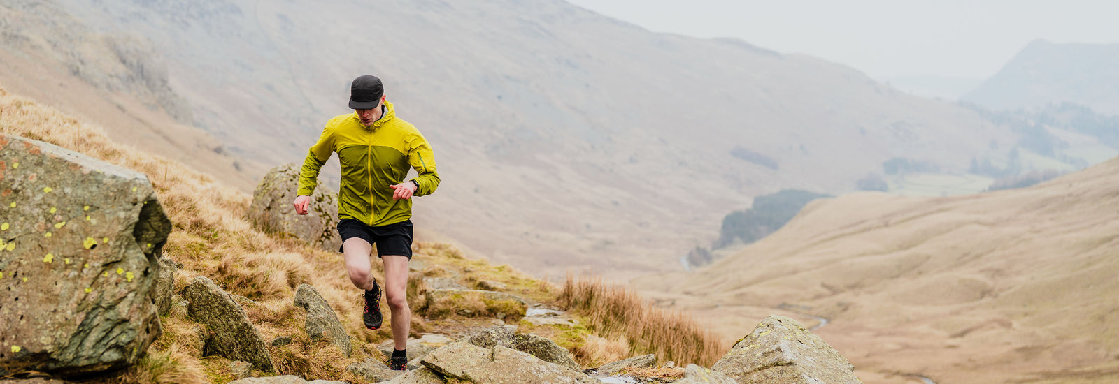 Trail runner in yellow jacket running across a rocky mountainside with expansive valleys beyond.