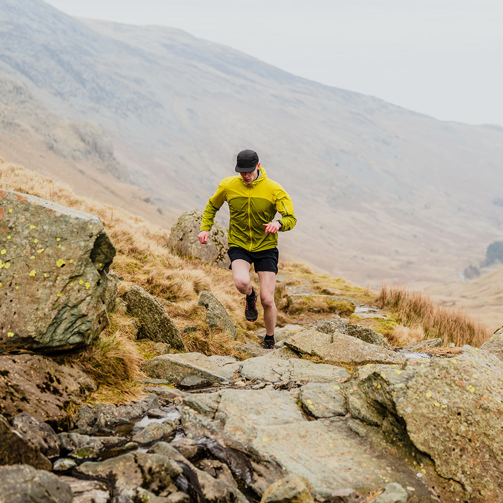 Trail runner in yellow jacket running over rocky mountain terrain with misty hills behind.
