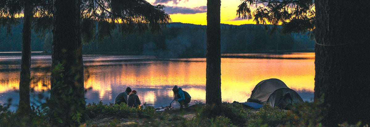 Campers beside a tent on a lake shore at sunset, surrounded by tall trees.